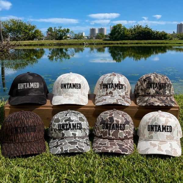 Collection of 'UNTAMED' branded hats in various designs on a wooden block by a lake.